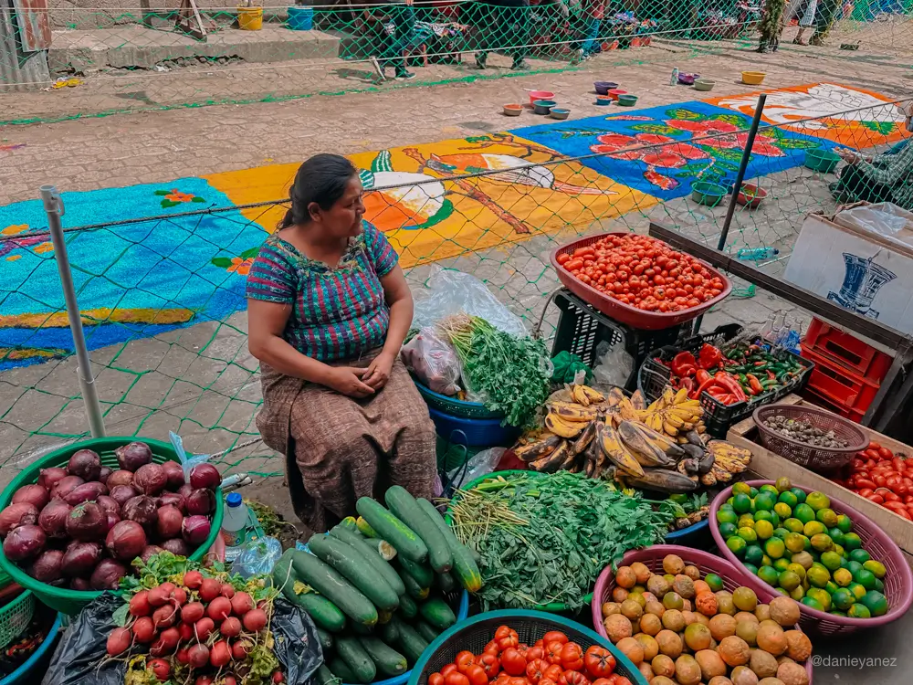 Mujer vendiendo fruta con alfombras de semana santa de fondo en Santiago Atitlán